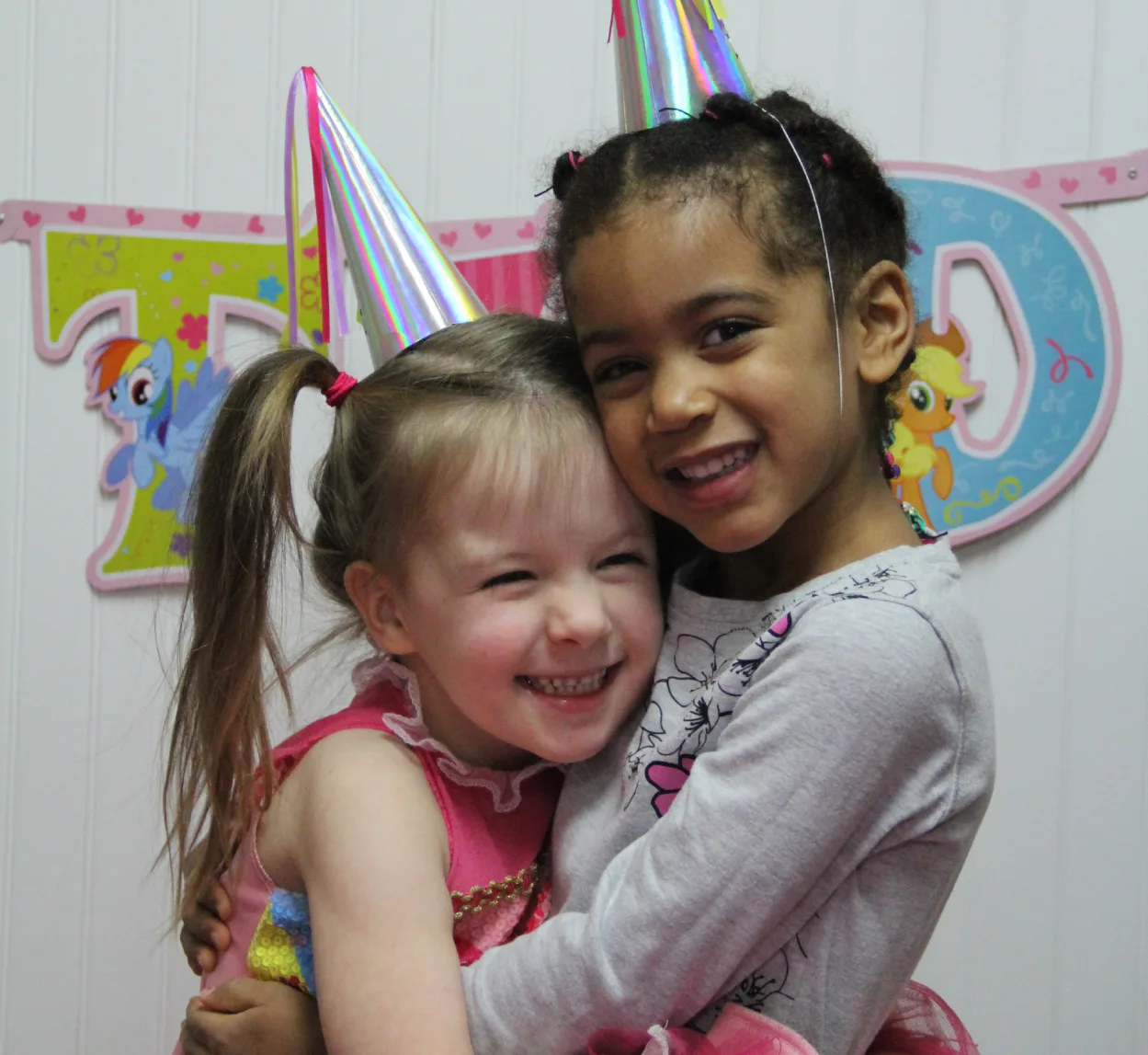 Deux fillettes coiffées de chapeaux de fête brillants se font un câlin en riant devant une banderole d'anniversaire