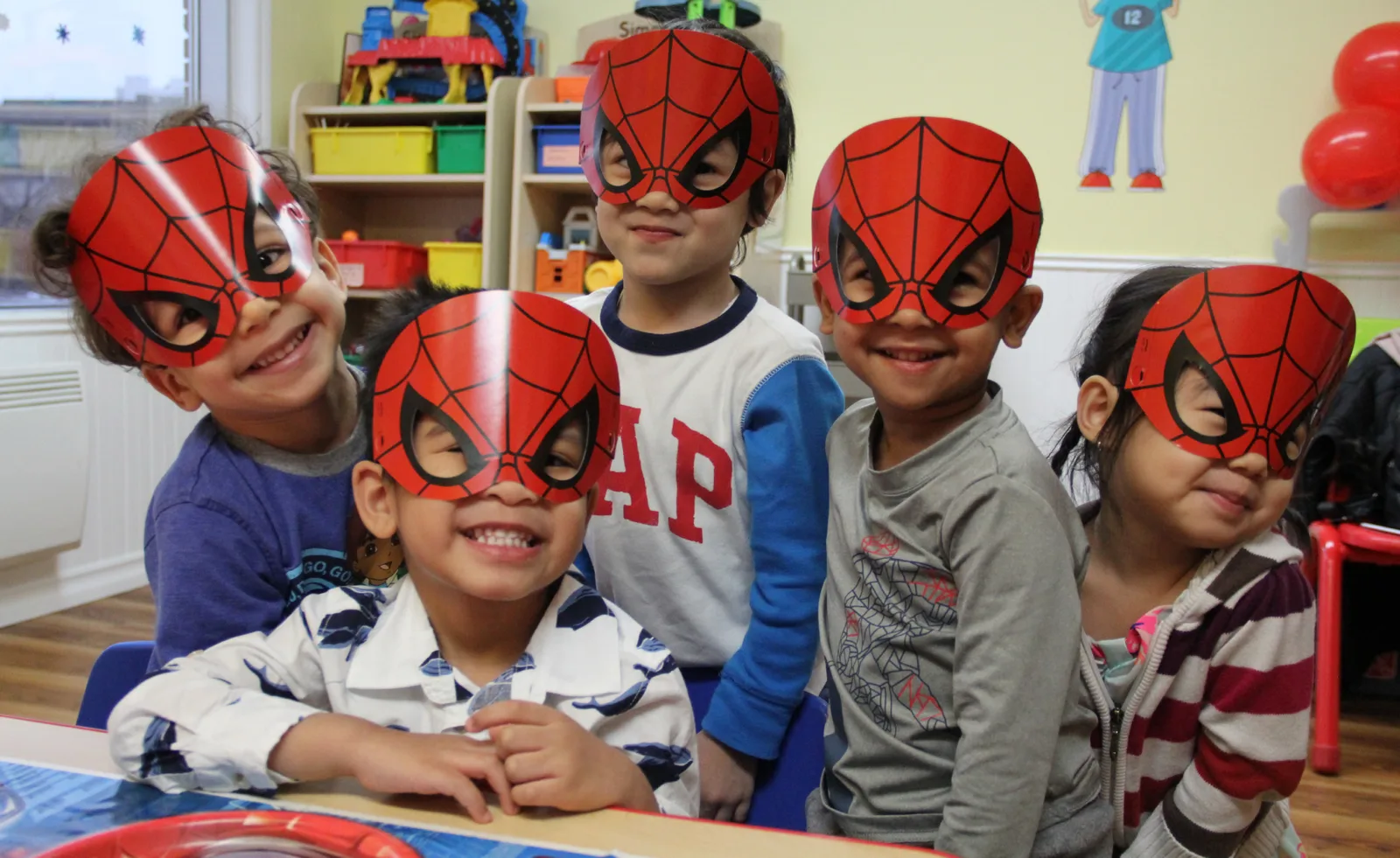 Cinq enfants souriants portant des masques de Spider-Man en papier réunis autour d'une table de fête à la garderie