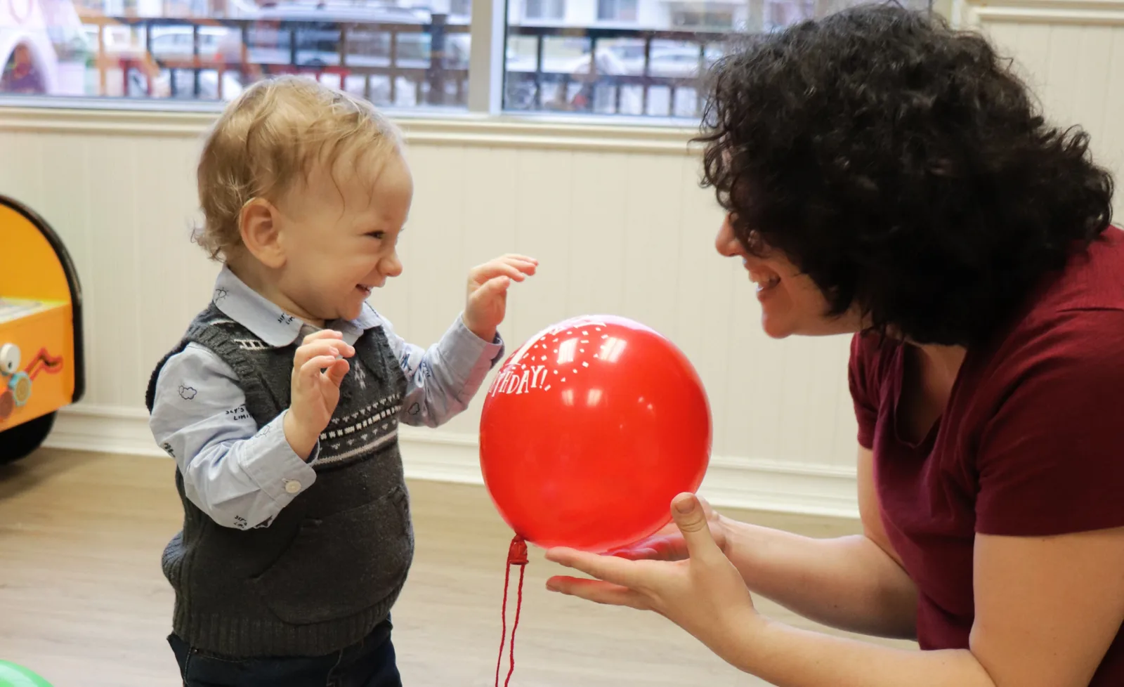 Un bambin tout souriant tend les mains vers un ballon de fête rouge que lui offre une éducatrice