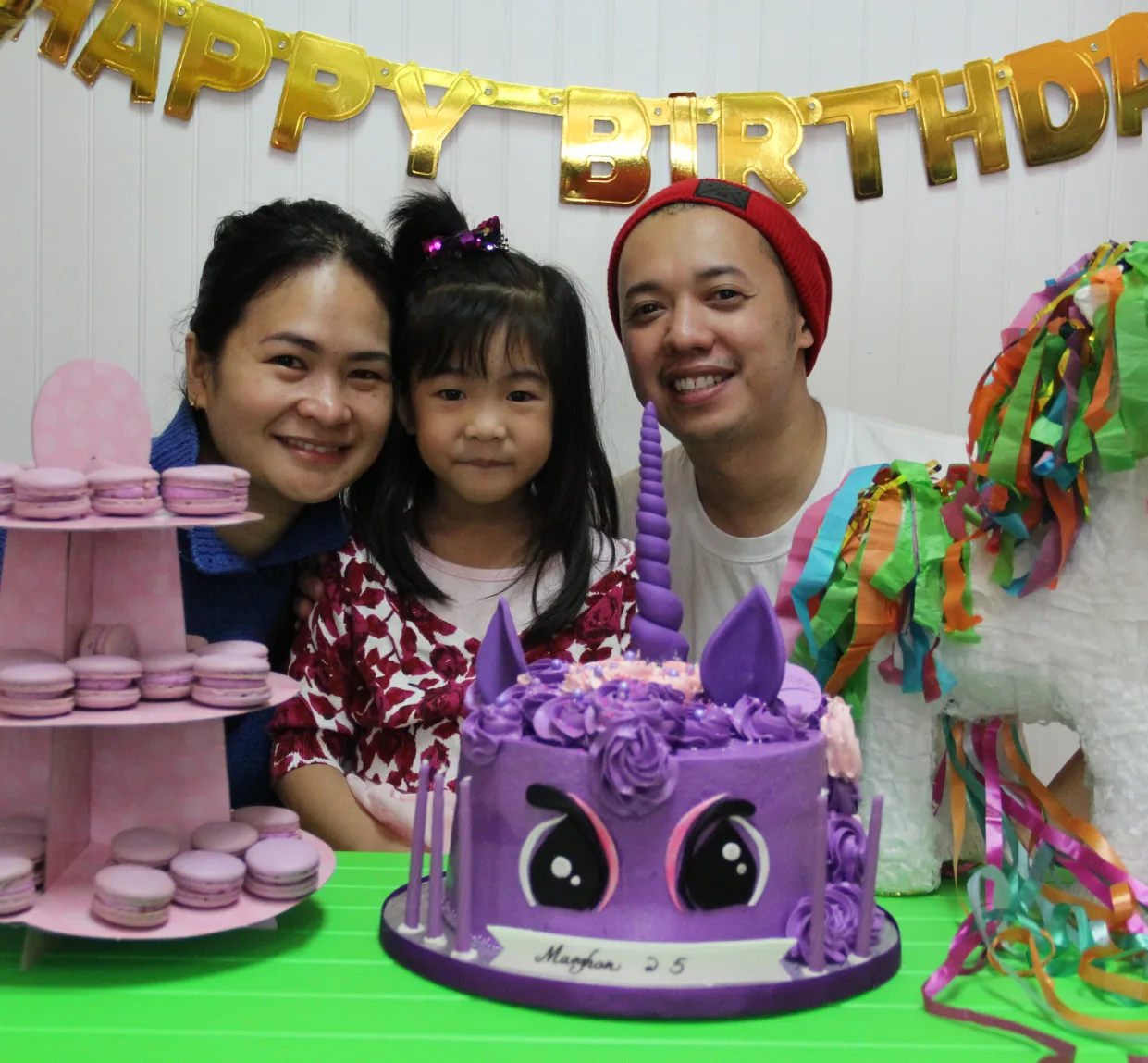 Une famille pose derrière un gâteau licorne mauve sous une banderole dorée de bonne fête