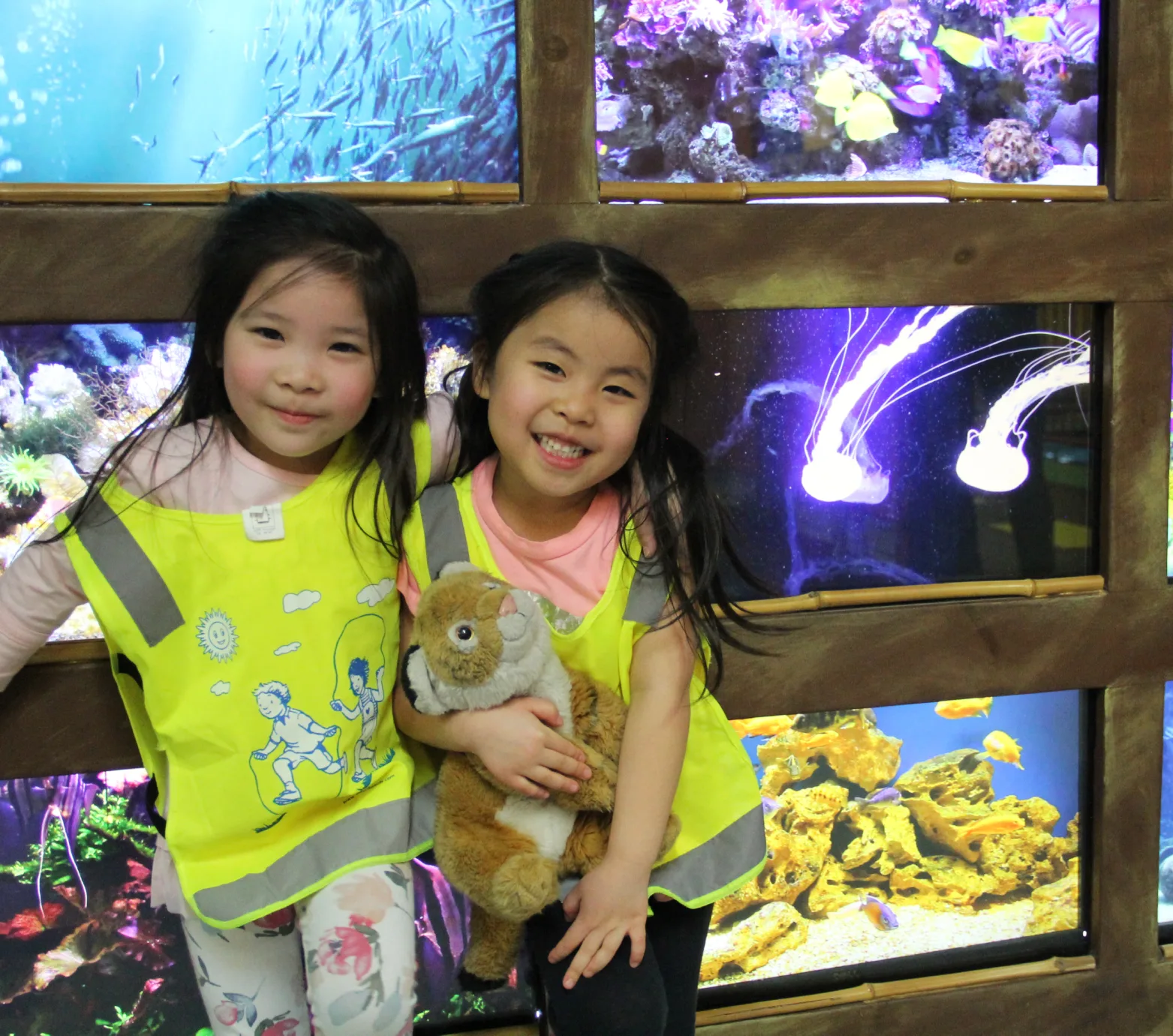 Deux fillettes souriantes en dossards jaunes posent avec un toutou devant les aquariums de méduses