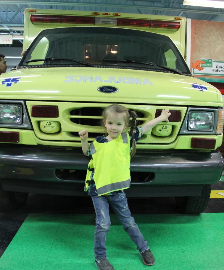 Un petit garçon en dossard jaune pose fièrement devant un camion d'ambulance