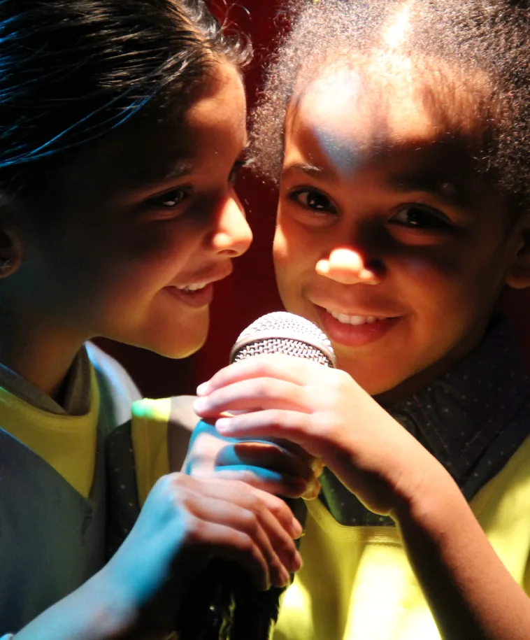 Deux enfants tout sourire partagent un micro sous un éclairage chaleureux de scène