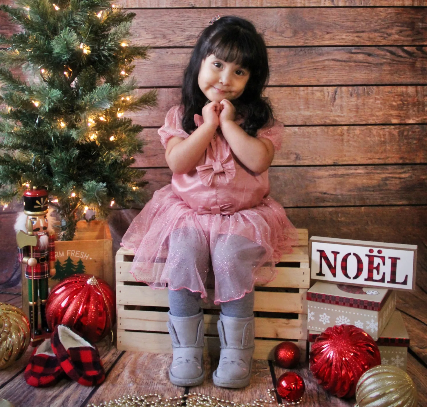 Fillette en rose assise sur une caisse de bois près du sapin illuminé et d'une affiche NOËL