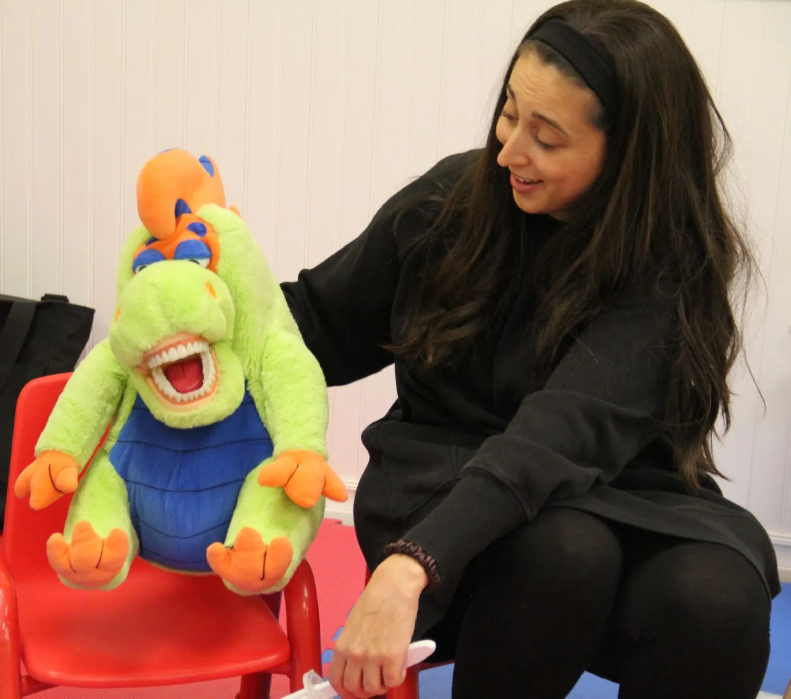 L'hygiéniste dentaire sourit à un dragon en peluche aux grandes dents assis sur une petite chaise rouge