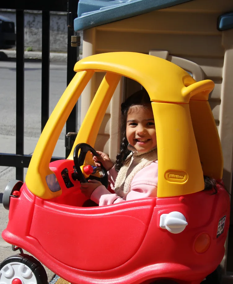 Fillette souriante au volant d'une petite voiture rouge et jaune dans la cour