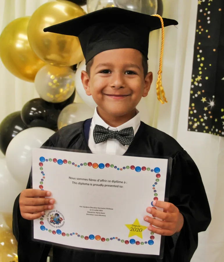 Un garçon en toge et mortier sourit fièrement avec son diplôme, près de ballons dorés.