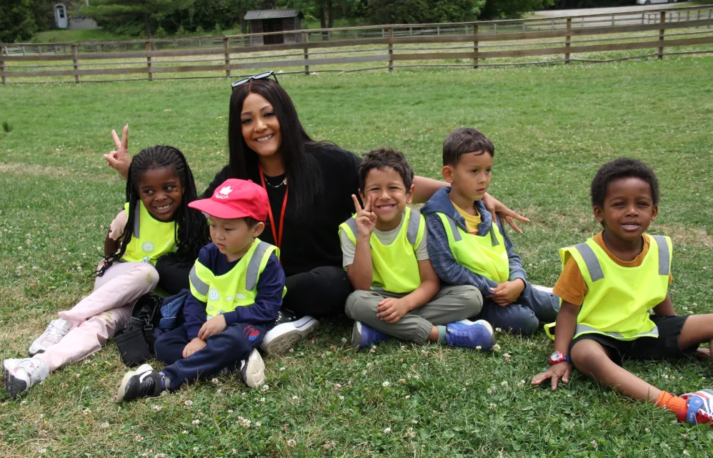 Une éducatrice et cinq enfants souriants en dossards jaunes assis dans l'herbe