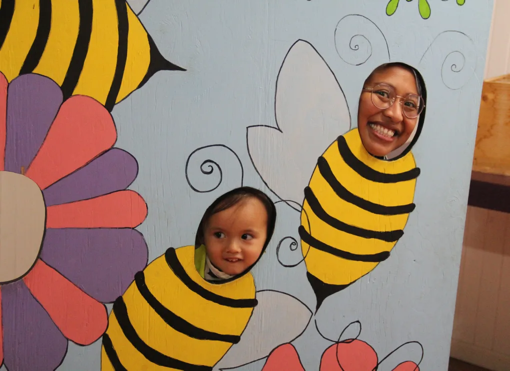 Un adulte et un enfant sourient à travers des découpes d'abeilles dans un panneau photo coloré