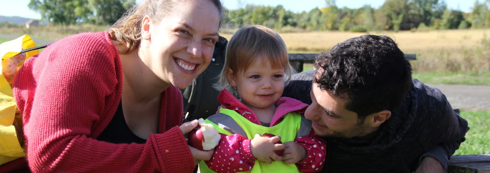Deux adultes souriants entourent une bambine qui tient une pomme dans un champ ensoleillé
