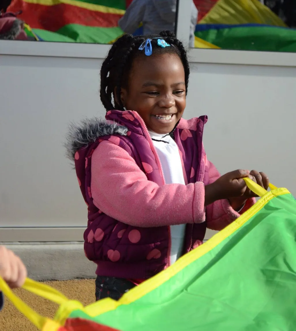 Une fillette souriante en manteau rose tient le bord du parachute coloré