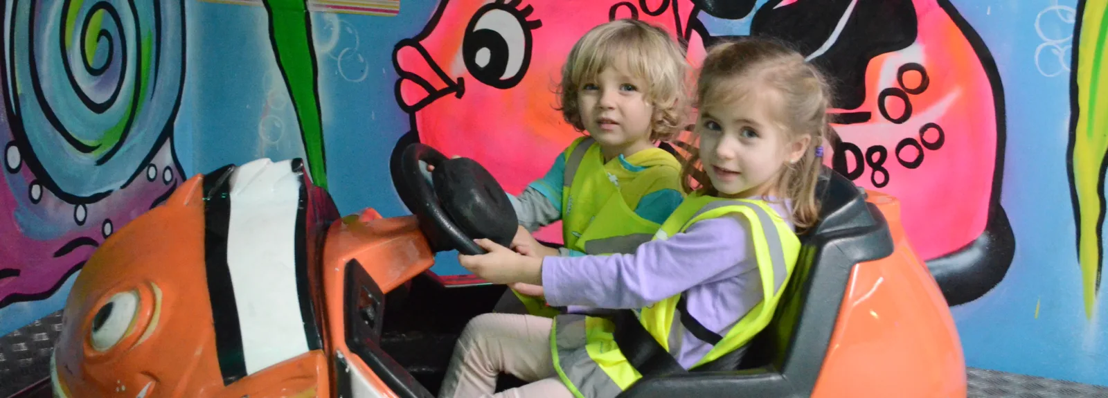 Deux enfants en dossards jaunes au volant d'un manège en forme de poisson orange devant une murale colorée