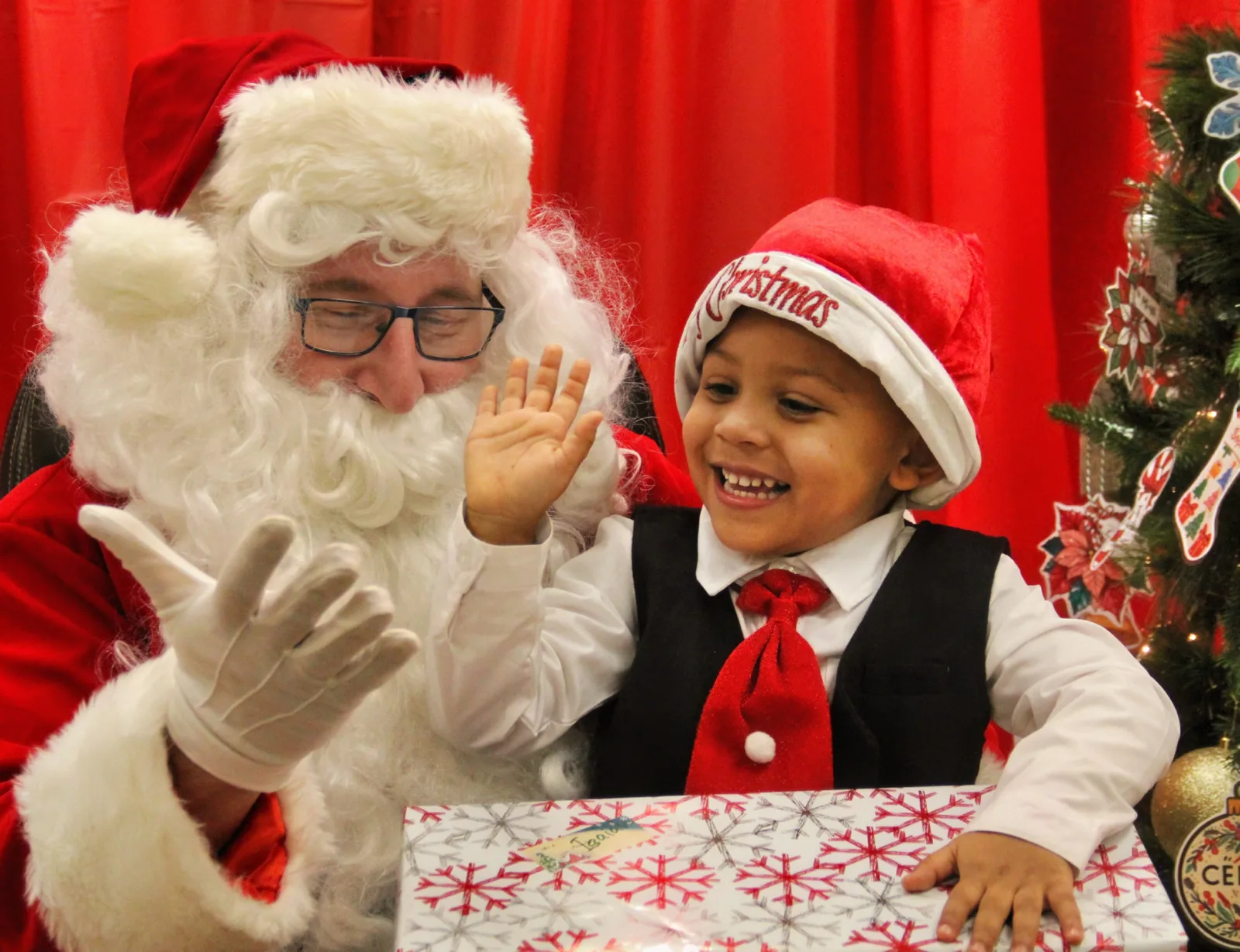 Père Noël qui salue un petit garçon rieur coiffé d'un chapeau de Noël, cadeau emballé devant lui