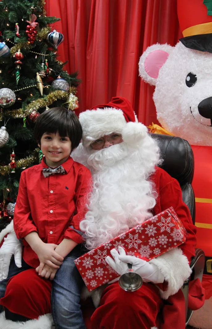 Garçon en chemise rouge et noeud papillon qui pose à côté du père Noël tenant un cadeau