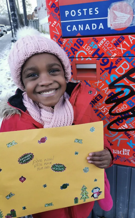Une fillette en tuque rose tient fièrement sa lettre décorée à côté d'une boîte aux lettres de Postes Canada