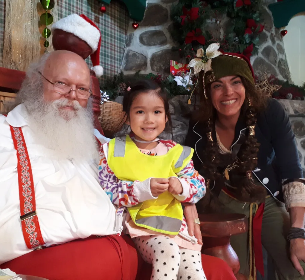 Une fillette en dossard jaune assise avec le père Noël et une lutine souriante