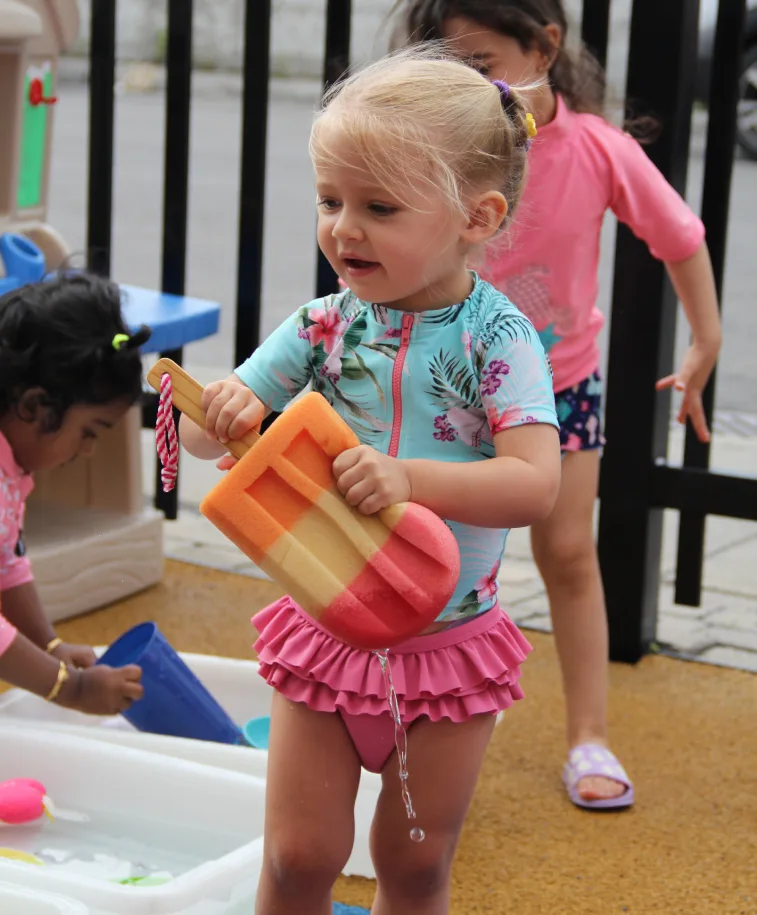 Fillette en maillot rose à froufrous qui tient des jouets en mousse près du bac d'eau