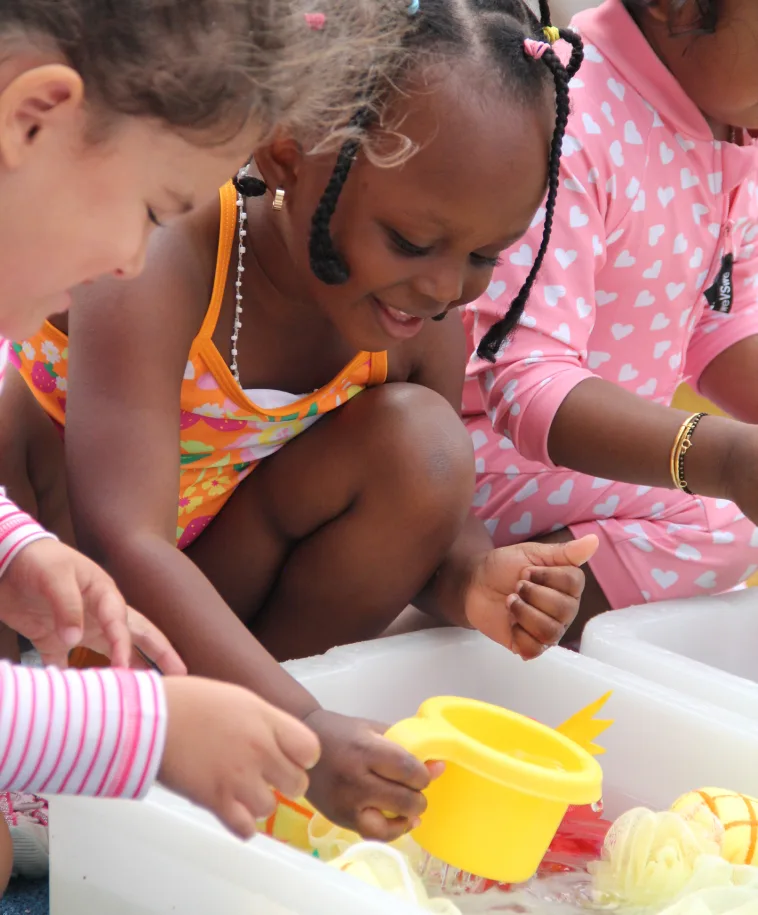Des enfants puisent de l'eau avec un seau jaune dans le bac sensoriel